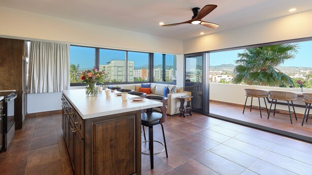 A modern kitchen with a large island, a dining area with a sofa and chairs, and a balcony with wicker furniture. - Beautiful condo located one block to Medano beach and The Cabo Marina (Cabo San Lucas)