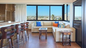A modern living room with a sofa, coffee table, and bar stools. A large window offers a view of the city. - Beautiful condo located one block to Medano beach and The Cabo Marina (Cabo San Lucas)