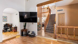 A spacious living area with a wooden floor, a flat-screen TV mounted on the wall, a staircase with wooden railings, and a door in the background. - Beautiful Santa Fe Oasis. Golf, hiking & biking paradise, 20 min drive to UofA. (Tucson)