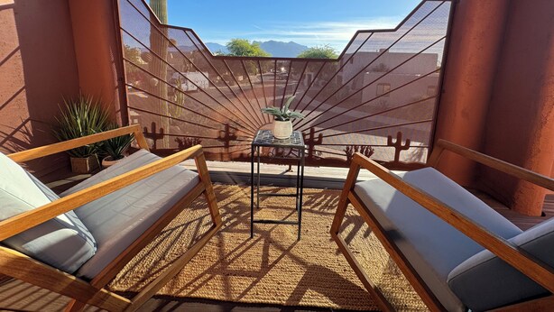 A sunlit outdoor seating area with wooden loungers and a woven mat, overlooking a cactus and mountains. - Beautiful Santa Fe Oasis. Golf, hiking & biking paradise, 20 min drive to UofA. (Tucson)