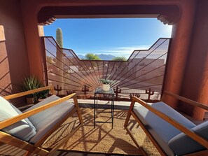 A sunlit outdoor seating area with wooden loungers and a woven mat, overlooking a cactus and mountains.