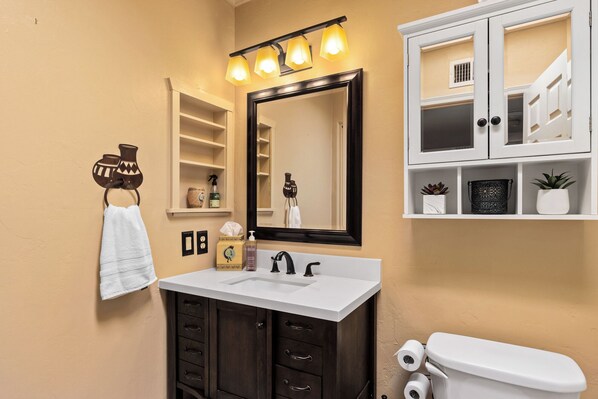 A bathroom with a white sink, dark wooden cabinets, a large mirror with lights, a towel rack, and a toilet.