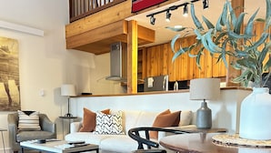 A modern living room with a sofa, coffee table, and dining table. There is a staircase leading to an upper level, and a potted plant is visible. - 2 Bed Remodeled Deluxe Mtn. Loft Minutes from Skiing JHMR (Wilson)