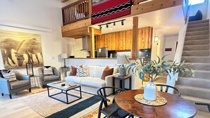 A spacious living area with a sofa, coffee table, and dining table. A staircase leads to an upper level with a balcony. - 2 Bed Remodeled Deluxe Mtn. Loft Minutes from Skiing JHMR (Wilson)