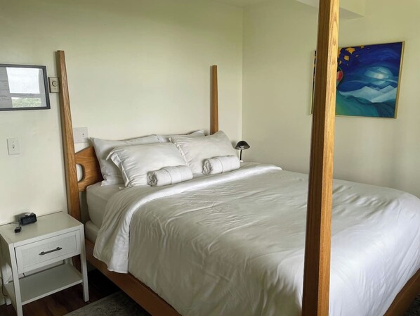 A neatly made bed with white linens, a wooden headboard, a bedside table with a lamp, and a painting on the wall.