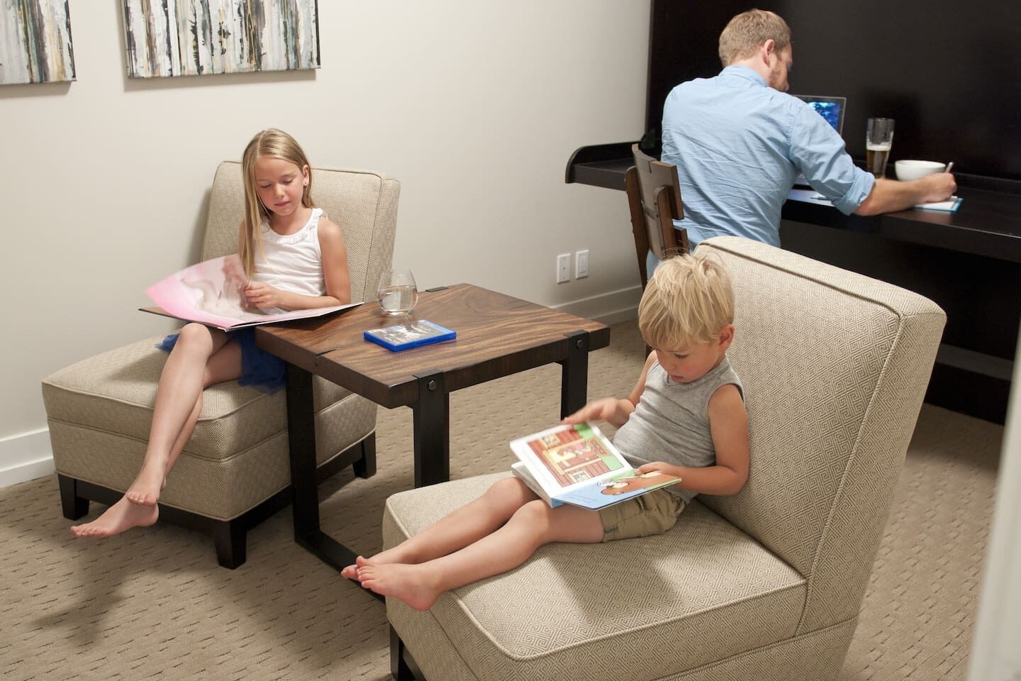 A living room with two children reading books, a man working at a desk, and a coffee table with a glass and a book.