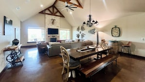 A spacious living area with a high ceiling, a fireplace, a large TV, and a dining table with benches. - Guadalupe River, Whitewater Amphitheater, New Braunfels, Canyon Lake, Gruene (New Braunfels)