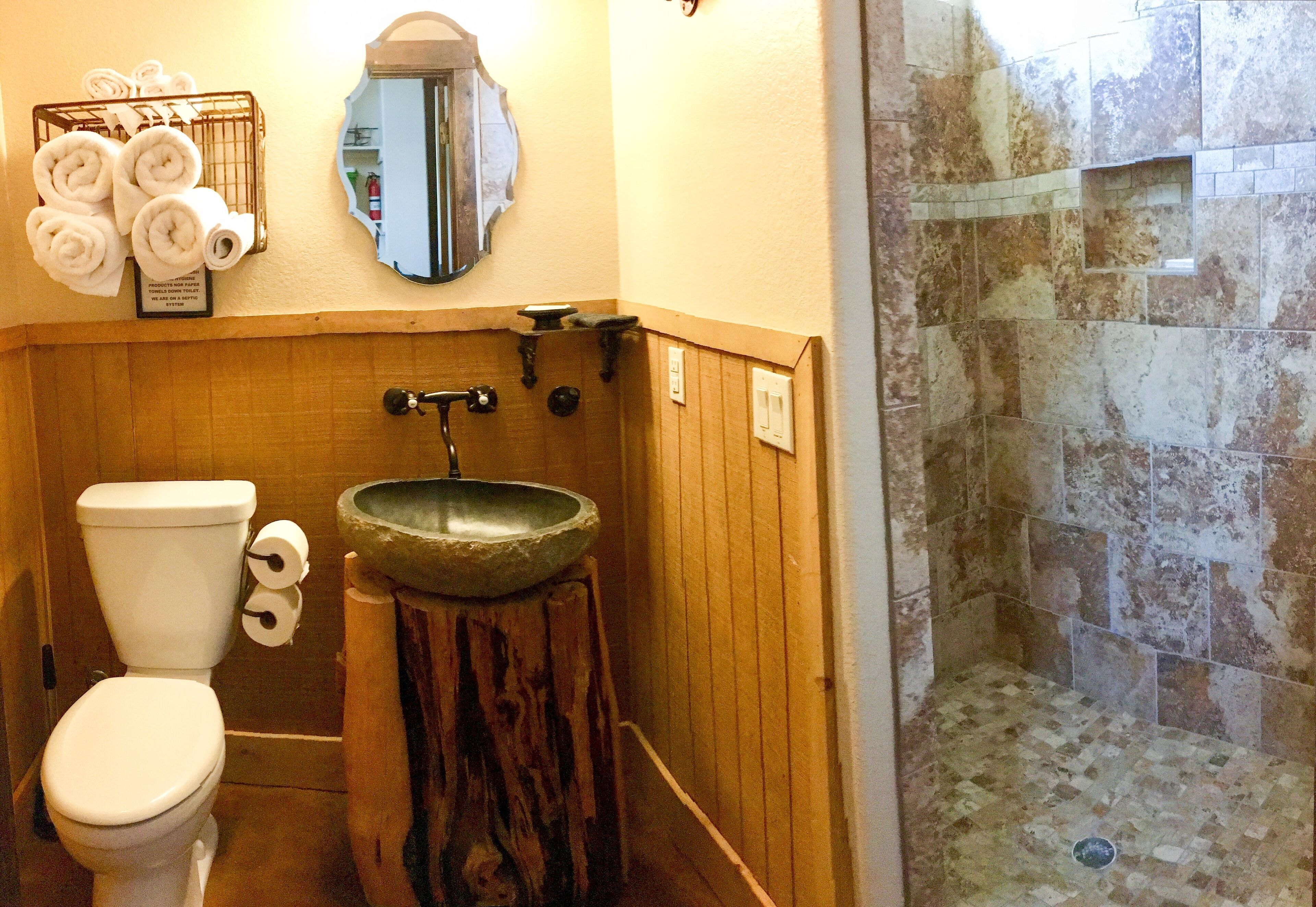 A bathroom with a toilet, a wooden vanity with a stone sink, a towel rack with towels, a mirror, and a shower area with stone tiles.