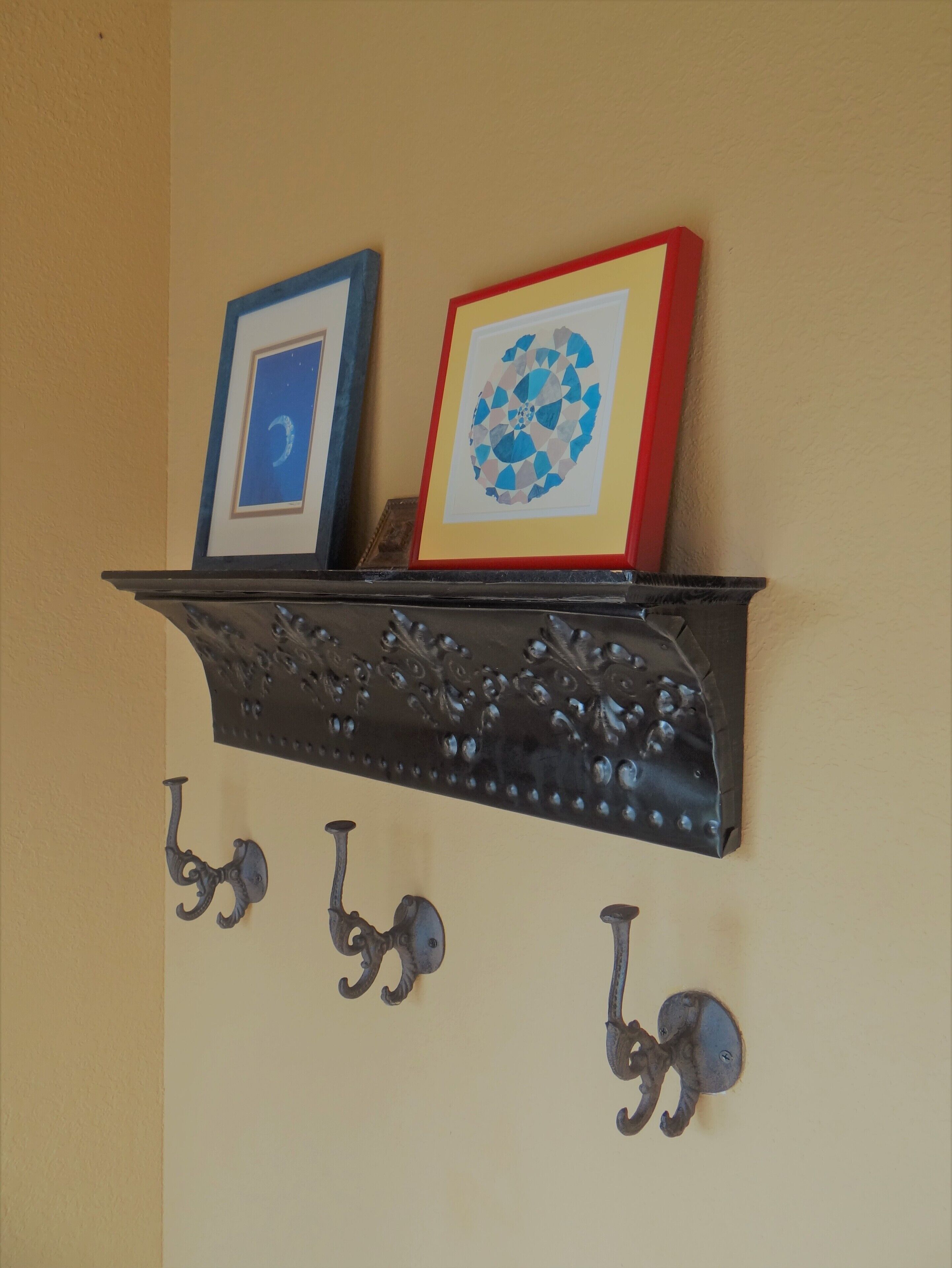 A black metal shelf with two framed artworks and three wall-mounted hooks.