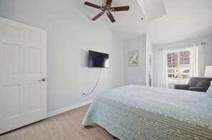Un dormitorio con una cama, ventilador de techo, televisor montado y ventana con cortinas.