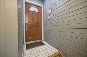 A wooden front door with a glass panel and a welcome sign, a tiled entrance mat, and a gray exterior wall.