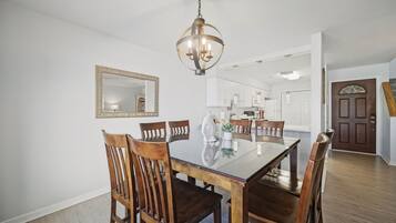 A dining area with a glass table, wooden chairs, a chandelier, and a kitchen in the background.