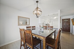 A dining area with a glass table, wooden chairs, a chandelier, and a kitchen in the background.