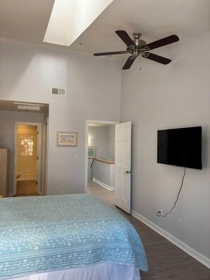 A bedroom with a bed, a ceiling fan, a TV mounted on the wall, and a door leading to another room.