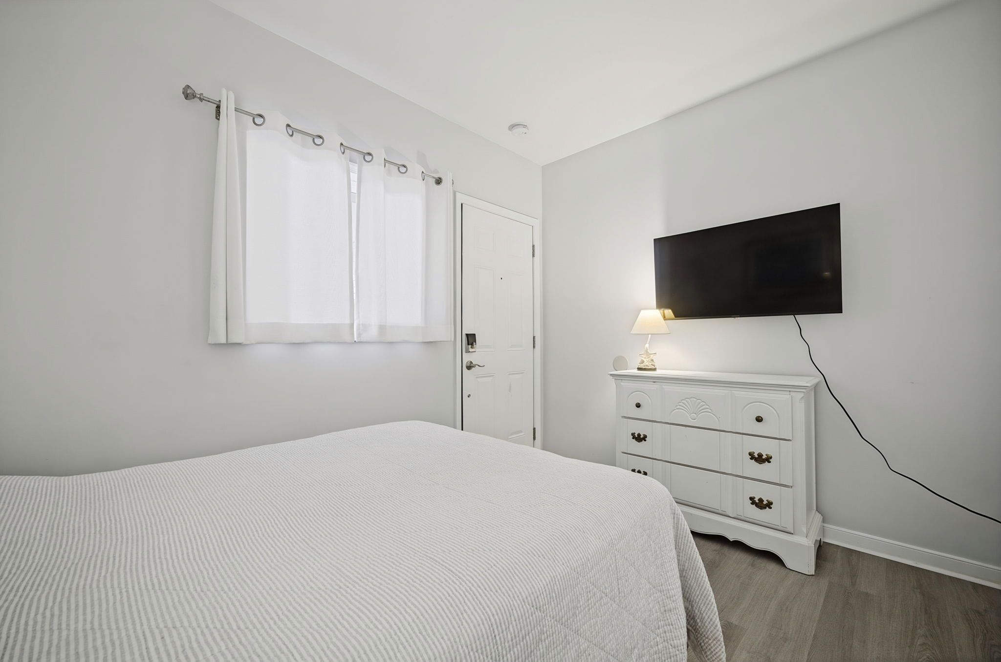 Un dormitorio con cama, cómoda, televisor montado en la pared y ventana con cortinas.