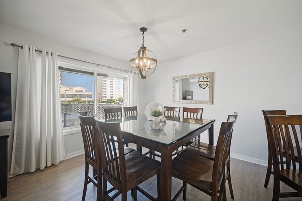 A dining area with a glass table, wooden chairs, a chandelier, a large mirror, and a view of buildings through a window.