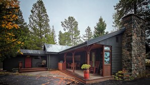 Una cabaña de troncos con chimenea de piedra, un porche cubierto con asientos y vistas al bosque circundante.