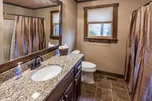 A bathroom with a granite countertop sink, a large mirror, a toilet, and a window with a view of trees.