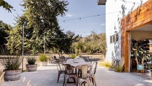 Un patio al aire libre con una mesa redonda y sillas, rodeado de plantas en macetas y un gran árbol.
