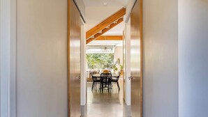 Un pasillo que conduce a un comedor con una mesa y sillas, vigas de madera en el techo y vistas al exterior con vegetación.