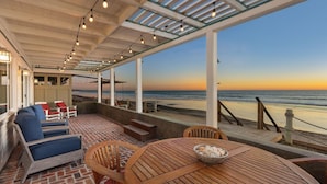A covered outdoor seating area with a view of the beach at sunset, featuring lounge chairs, a wooden table, and string lights. - OCEANFRONT CHIC LUXURY HOME (Capistrano Beach)