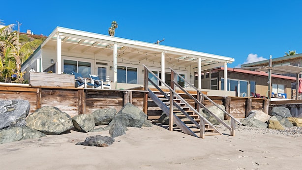 A beachfront house with a covered porch, a staircase leading to a deck, and a sandy beach in the foreground. - OCEANFRONT CHIC LUXURY HOME (Capistrano Beach)