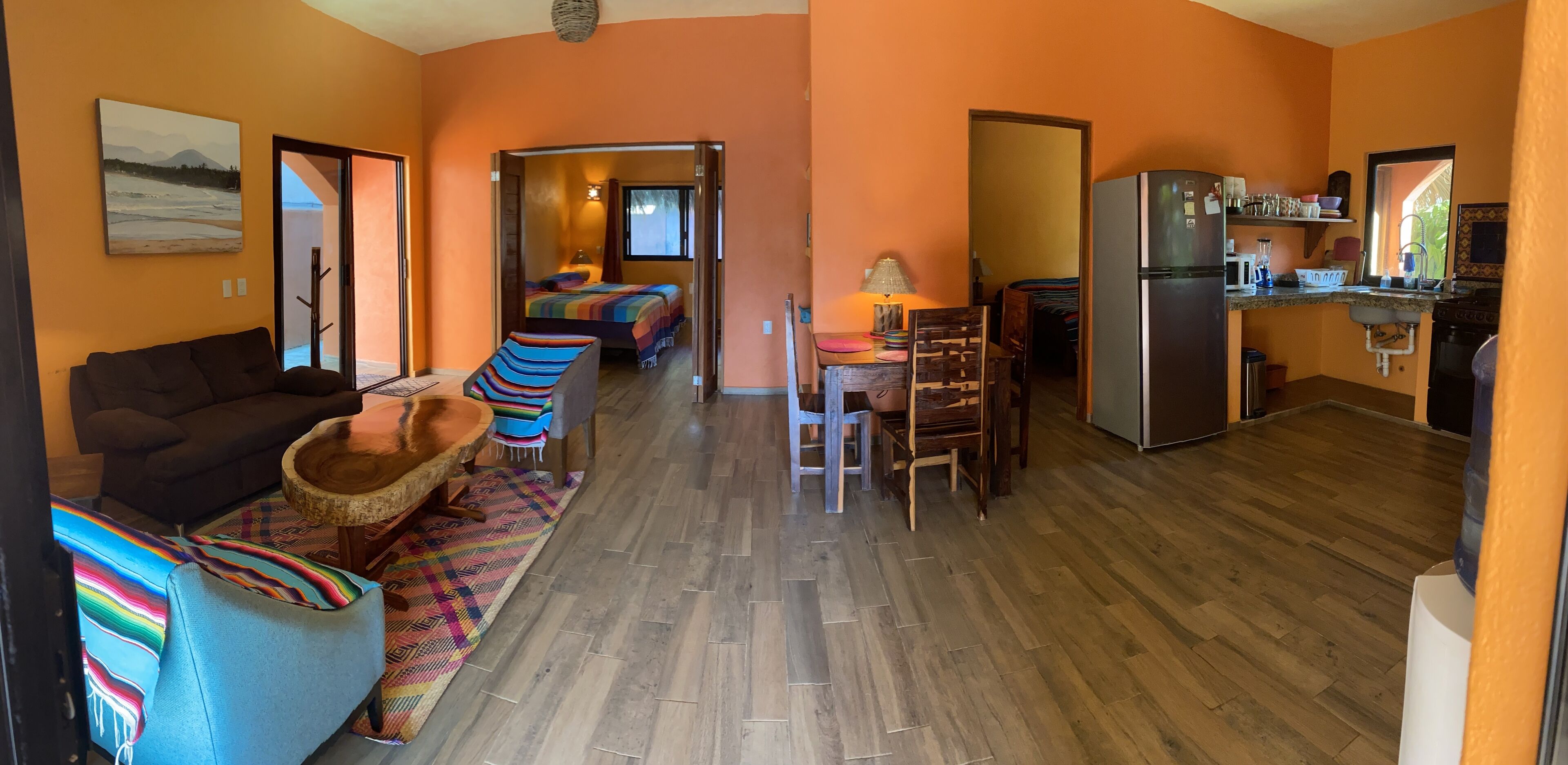 Un espacio de estar acogedor con un sofá, un tapete colorido y una mesa de centro de madera. En el fondo, se ve una cama con una manta de colores del arcoíris.