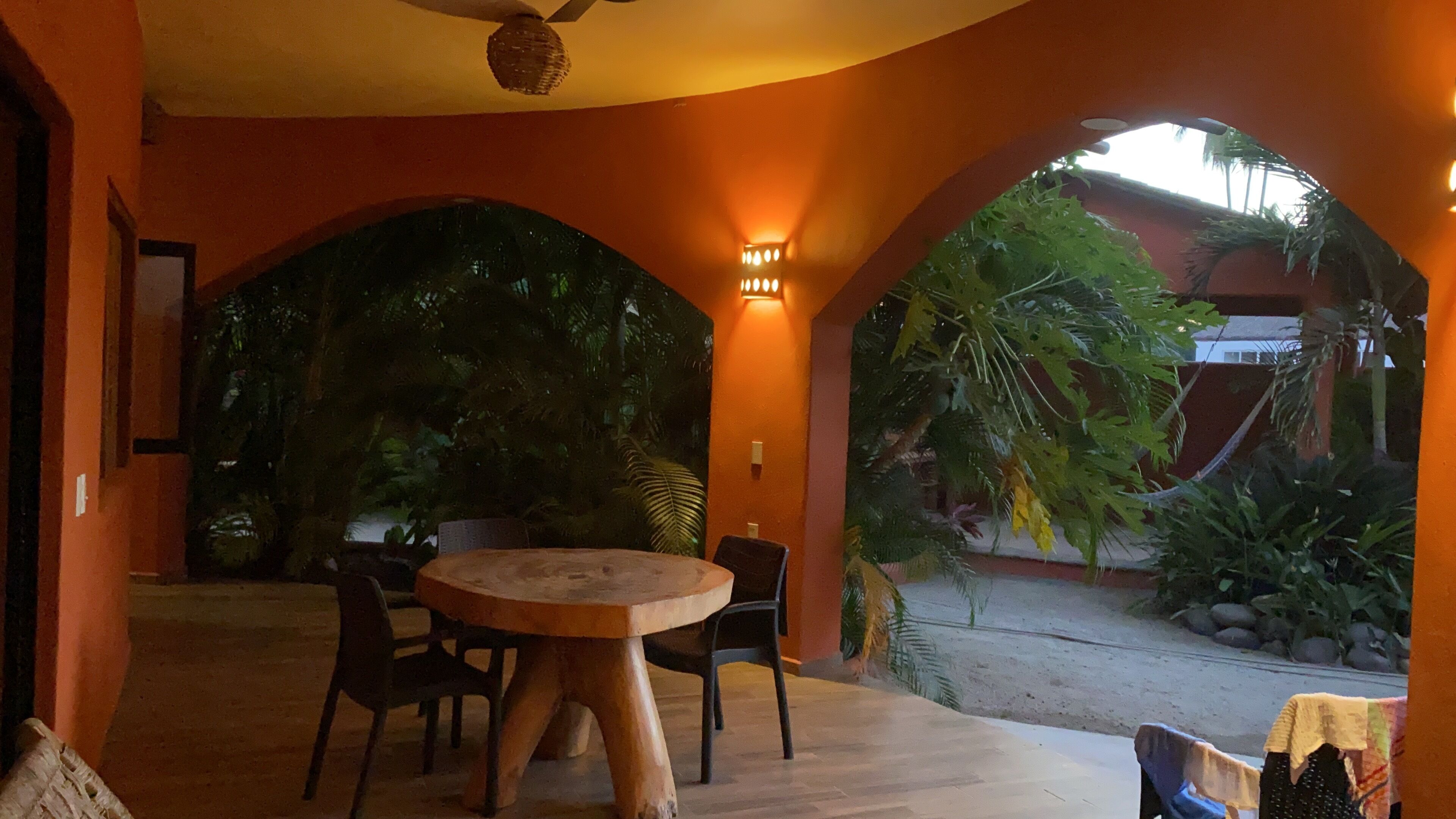 Ein Essbereich mit einem runden Holztisch und Stühlen, umgeben von orangefarbenen Wänden und Blick auf einen Garten mit tropischen Pflanzen.