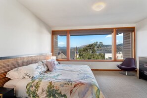 Ein Schlafzimmer mit einem großen Fenster, einem Bett mit floralem Bettbezug, einem Holz-Kopfteil und einem violetten Sessel.