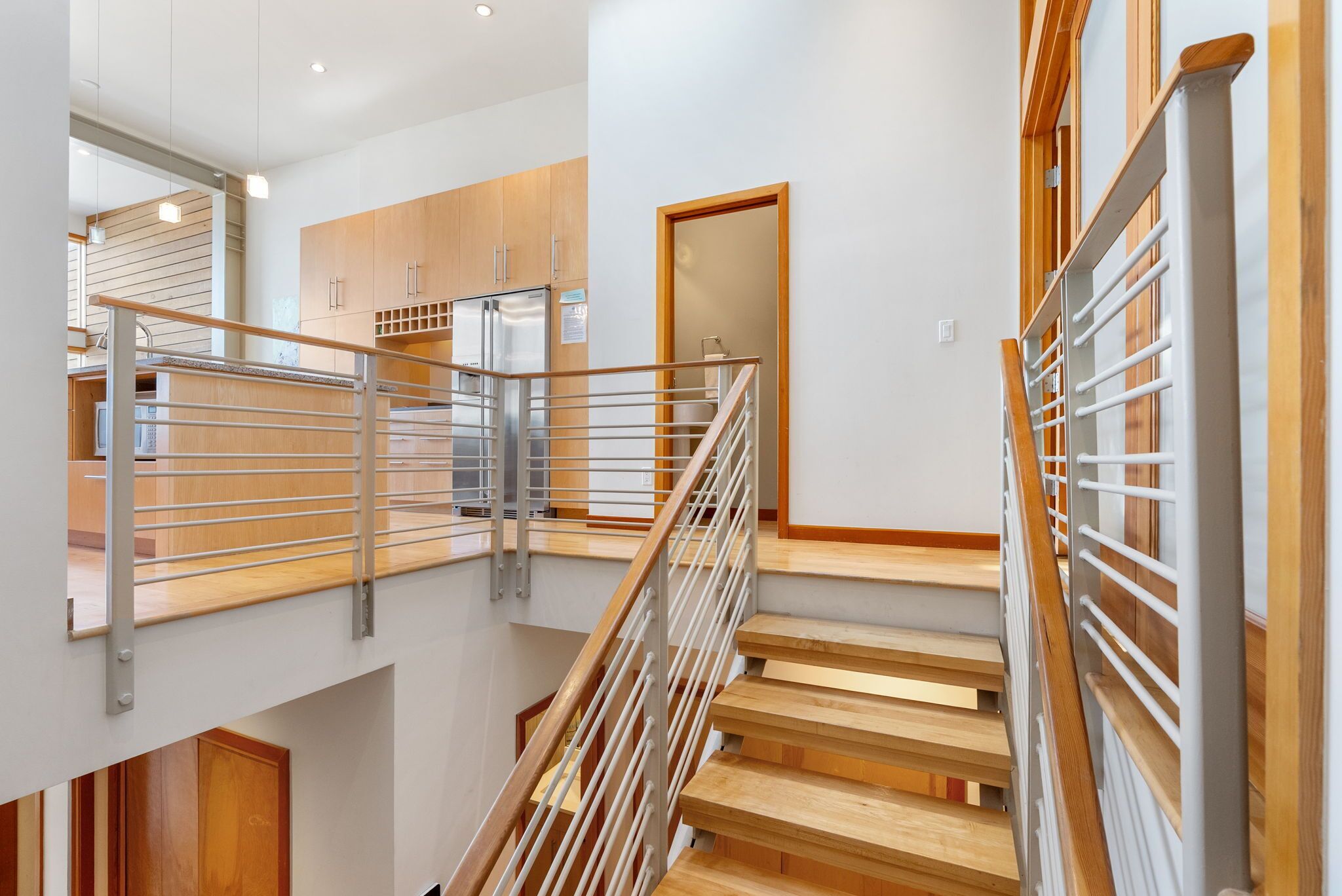 Un intérieur moderne avec un escalier en bois, des rampes en métal et un coin cuisine doté de meubles en bois et d’un réfrigérateur.