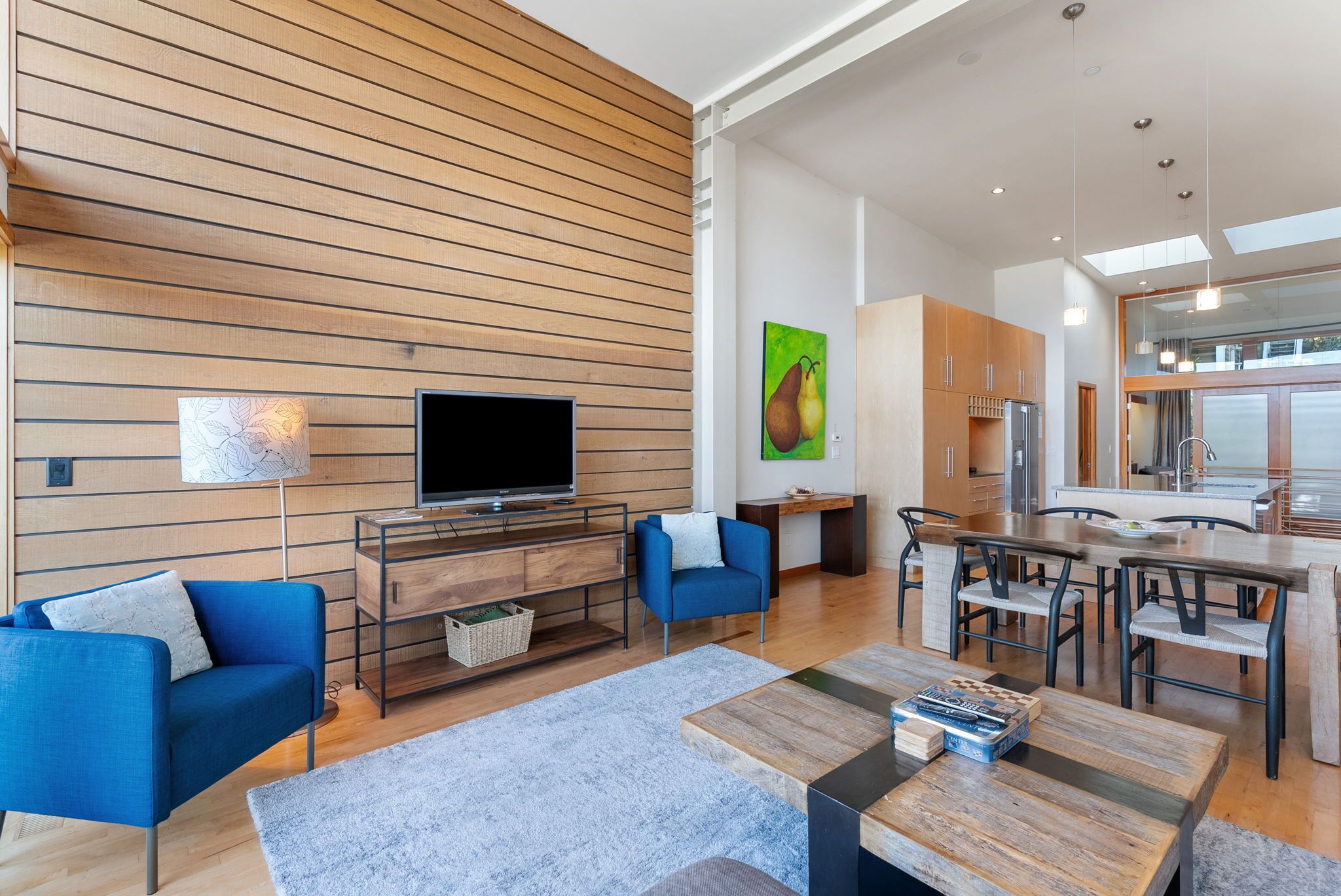 Un salon moderne doté d’un téléviseur à écran plat, de canapés bleus, d’une table basse en bois et d’un coin cuisine avec des meubles en bois et une table à manger.