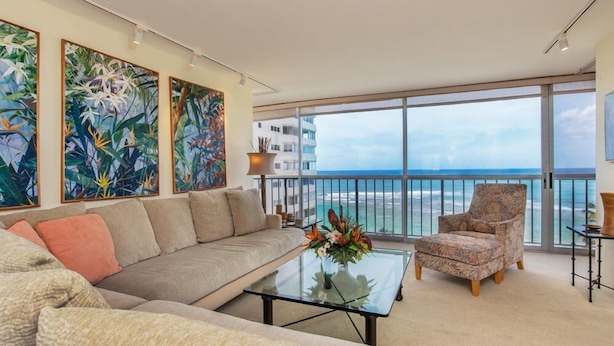A spacious living room with a large sofa, a coffee table, and a chair. There are three large paintings on the wall, and a balcony with a view of the ocean.