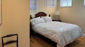 Un dormitorio con cama de madera, ropa de cama blanca, una mesita de noche con lámpara y dos ventanas con persianas.