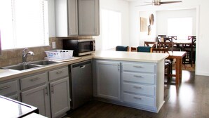 Una cocina con armarios grises, microondas y lavavajillas. Hay un comedor con sillas de madera y una mesa.