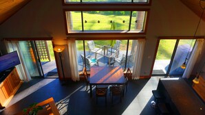 A dining area with a table and chairs, a television, and a view of a grassy yard through large windows. - Contemporary Oceanfront Cottage with Private Beaches, Trails, and Gardens (Sooke)