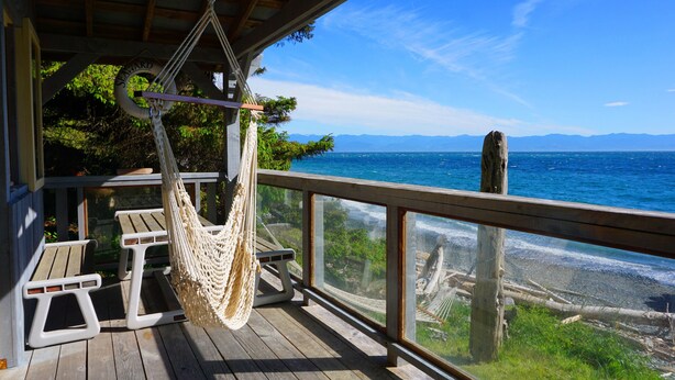 A wooden deck with a hammock, picnic table, and a view of the ocean and mountains. - Contemporary Oceanfront Cottage with Private Beaches, Trails, and Gardens (Sooke)
