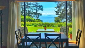 A dining area with a table and chairs, large windows with curtains, and a view of a green landscape and the sea. - Contemporary Oceanfront Cottage with Private Beaches, Trails, and Gardens (Sooke)