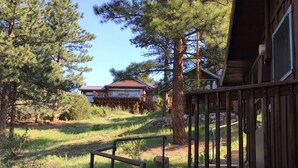 A wooden cabin with a deck, surrounded by pine trees and a grassy area. - In the center of town with views of Longs Peak (Estes Park)