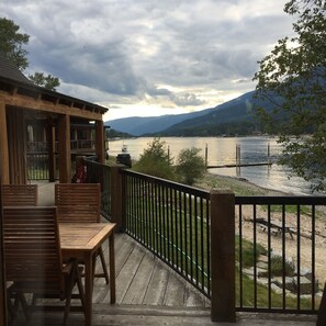 Terraza de madera con mesa y sillas, con vista a un lago y montañas al fondo.