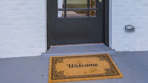 Una puerta negra con un panel de vidrio, dos luces fijadas a la pared, un felpudo con la palabra 'Welcome' y una pared blanca con textura.