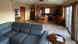 A spacious living room with a large gray sectional sofa, a wooden coffee table, and a small round table with a remote control. A kitchen with a central island and bar stools is visible in the background. - 1 BR guest house overlooking a beautiful vineyard  (oliver)