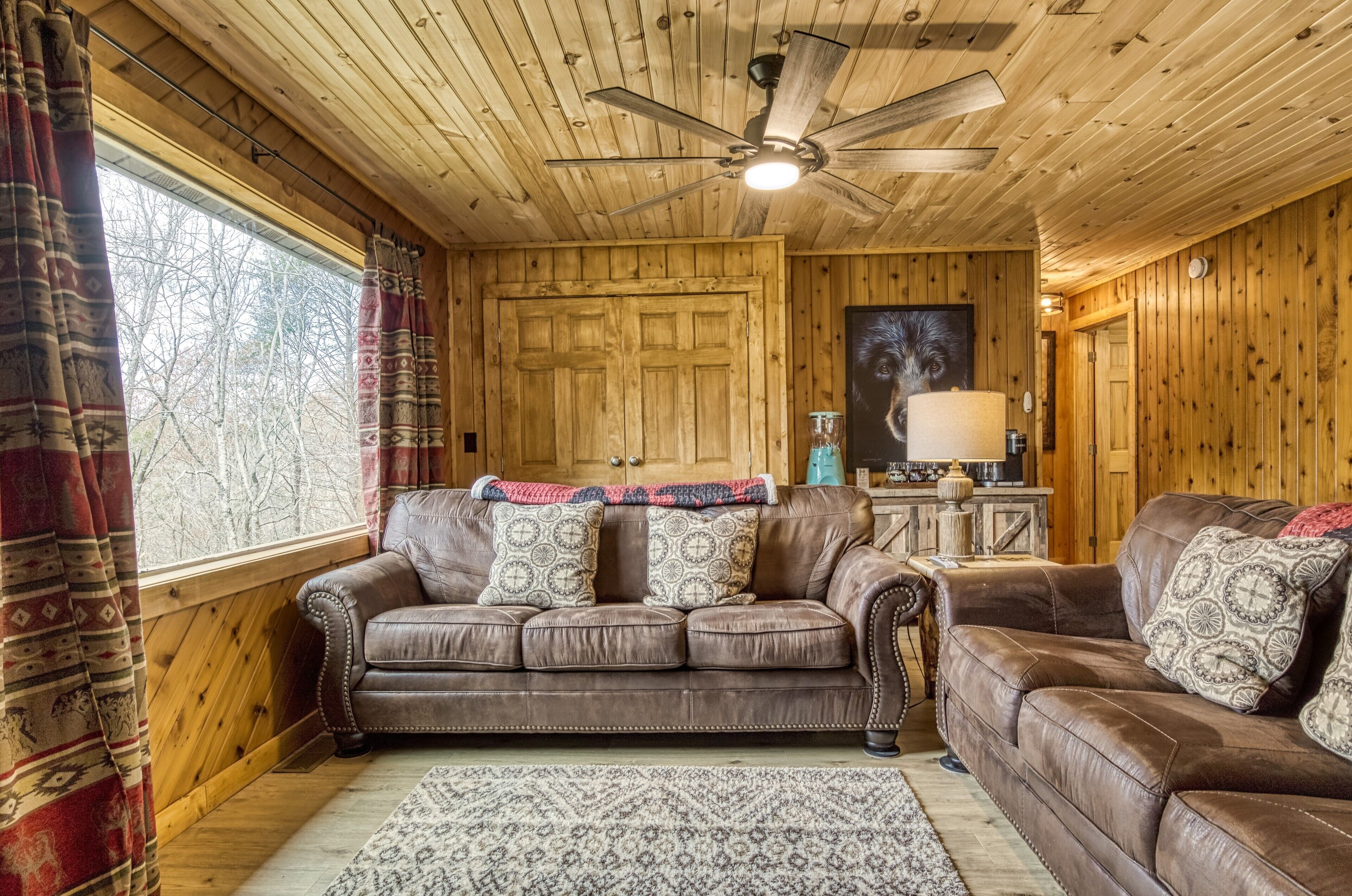 A cozy cabin interior with a wooden ceiling, a ceiling fan, a large window with curtains, and two brown leather sofas with patterned cushions.