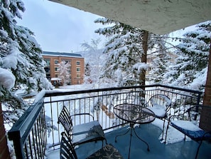 A balcony with a metal table and two chairs, covered in snow, overlooking a building with snow-covered trees.