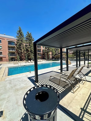 Zona de piscina con sillones reclinables, un espacio de estar cubierto y vistas a un edificio con balcones.