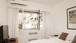 Hotellihuone, jossa on sänky, työpöytä kannettavalla tietokoneella, ilmastointilaite, ikkuna verhoilla ja seinällä oleva kehystetty kuva.