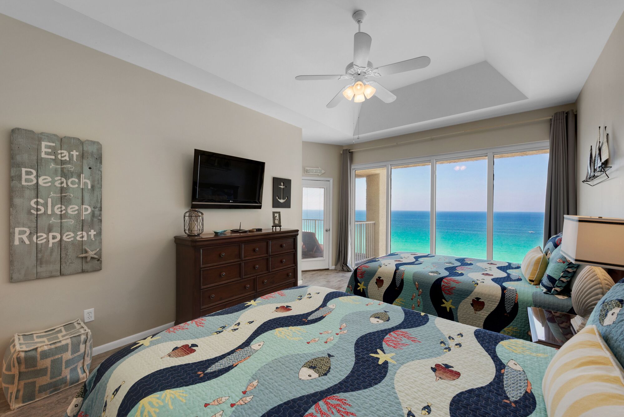 Een slaapkamer met een groot raam met uitzicht op de oceaan, een plafondventilator en een bed met een sprei.