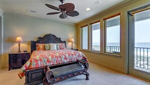 Un dormitorio con una cama grande, un ventilador de techo, dos lámparas de noche, una mesita de noche y vistas a la playa a través de amplias ventanas.