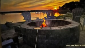 A stone fire pit with a fire, surrounded by Adirondack chairs, overlooking a body of water at sunset.