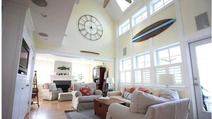 A spacious living room with a high ceiling, a large clock, and a surfboard decoration. - Harbour Island Coastal Home, shoulder season mid-week specials! Booking 2027 (Narragansett)