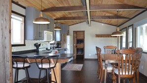 A spacious kitchen with wooden beams, a dining area with wooden chairs, and a view through an open door. - Controlled Chaos Basecamp - centered between Jackson & Clark County Trail System (Merrillan)